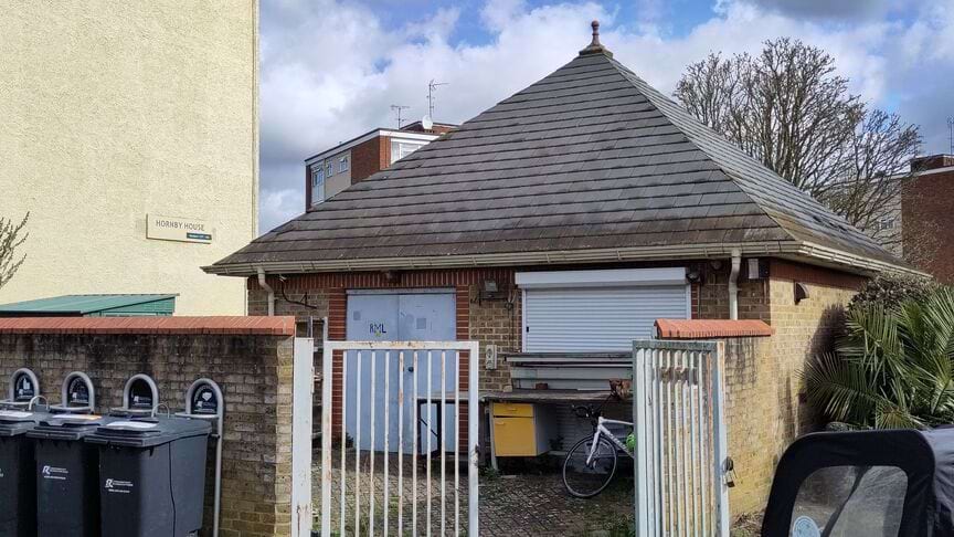 The front of the little house with An estate on the background, recycle bins can be seen to the left in front of little house.